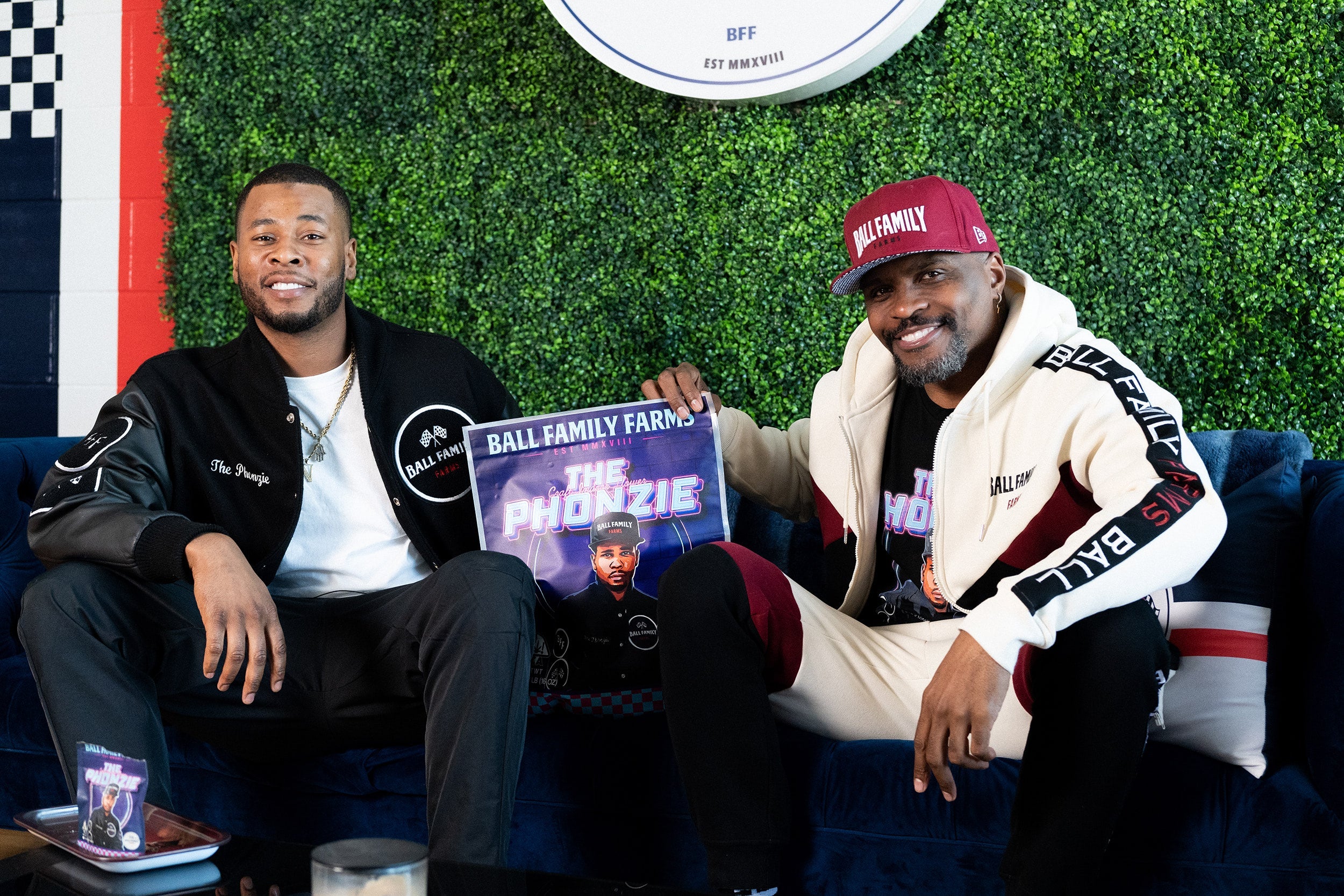 CEO Chris Ball and actor/musician J. Alphonse Nicholson sitting on a couch, displaying a poster of their cannabis strain.
