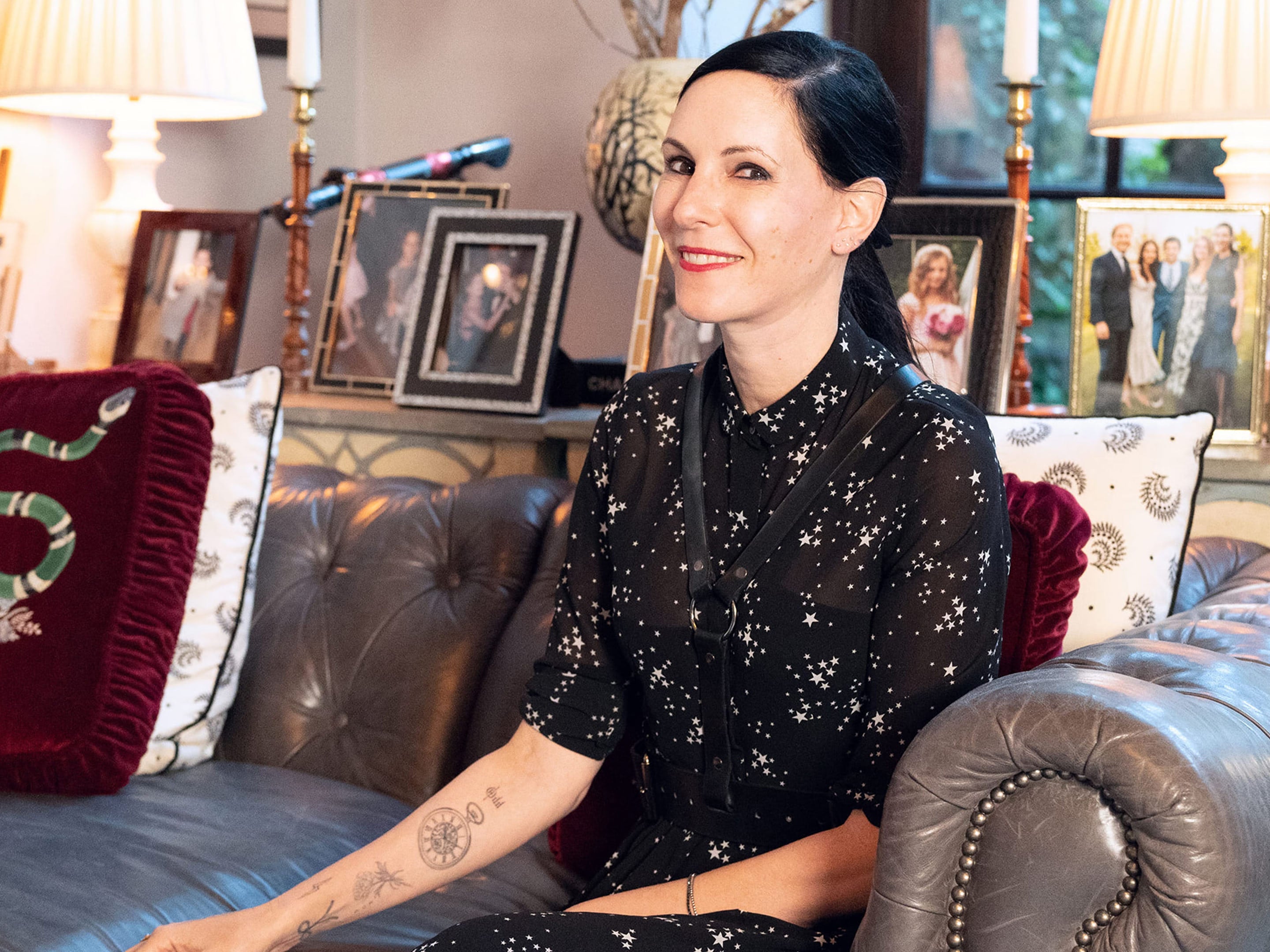Jill Kargman, actress and comedian, sitting on her couch at home with framed photos behind her.