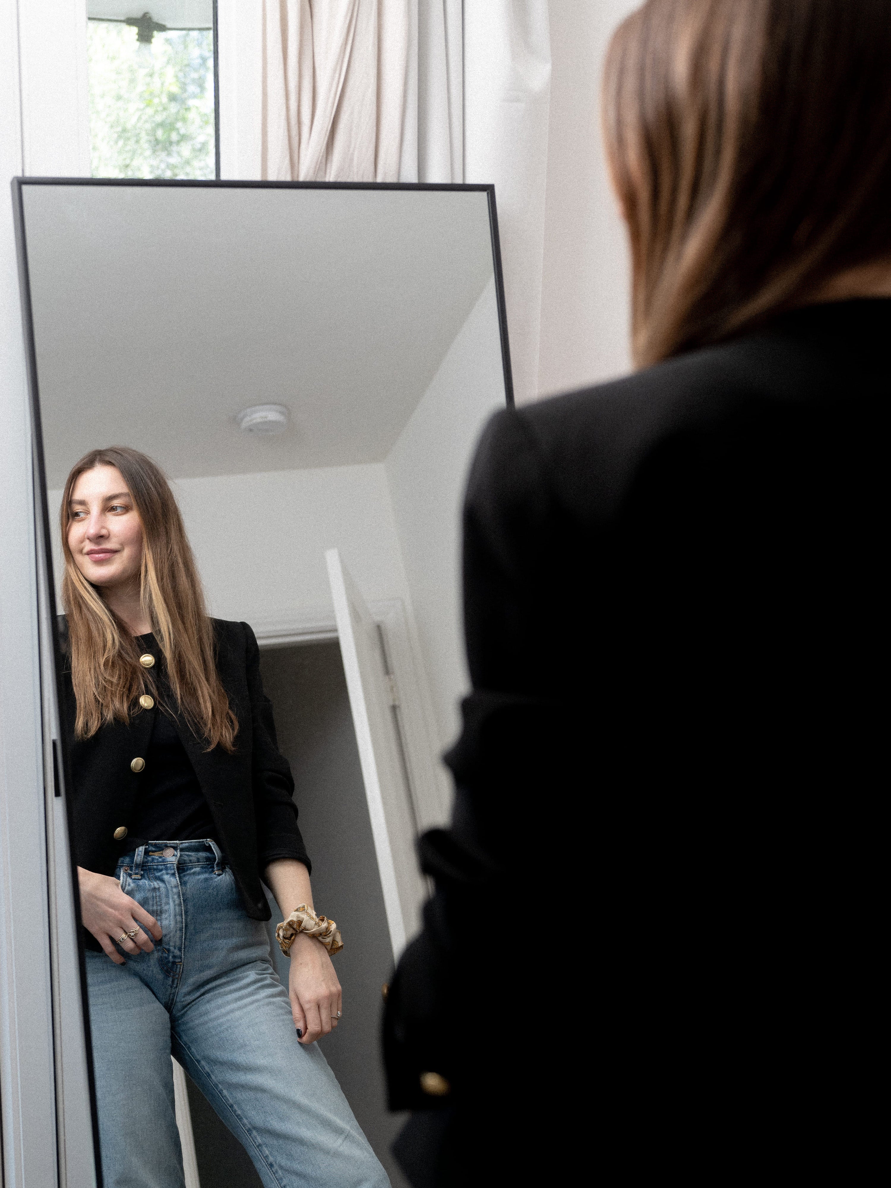 Over-the-shoulder view of Allison Bornstein in jeans and a black jacket, standing in front of a mirror.
