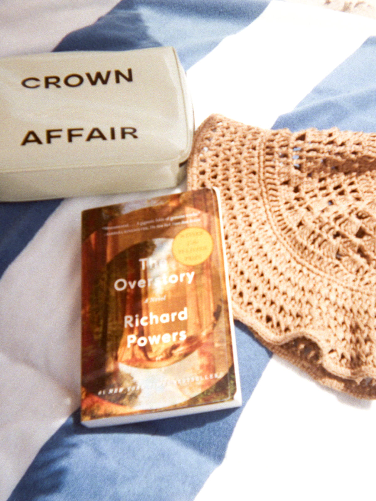 Dianna Cohen's Crown Affair bag, The Overstay book by Richard Powers and a hat on on a towel in the sun