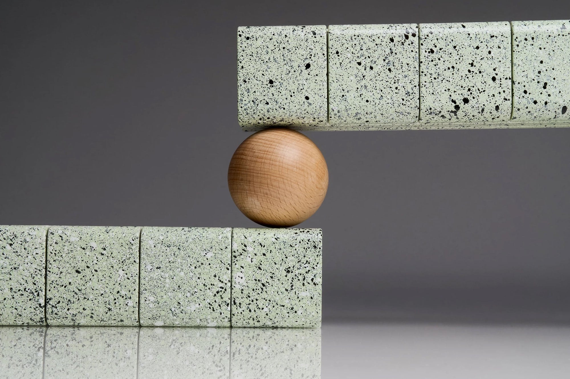 Speckled green AndAgain 7-day hand-painted pill cases balanced around a wooden sphere in a minimalist studio composition.