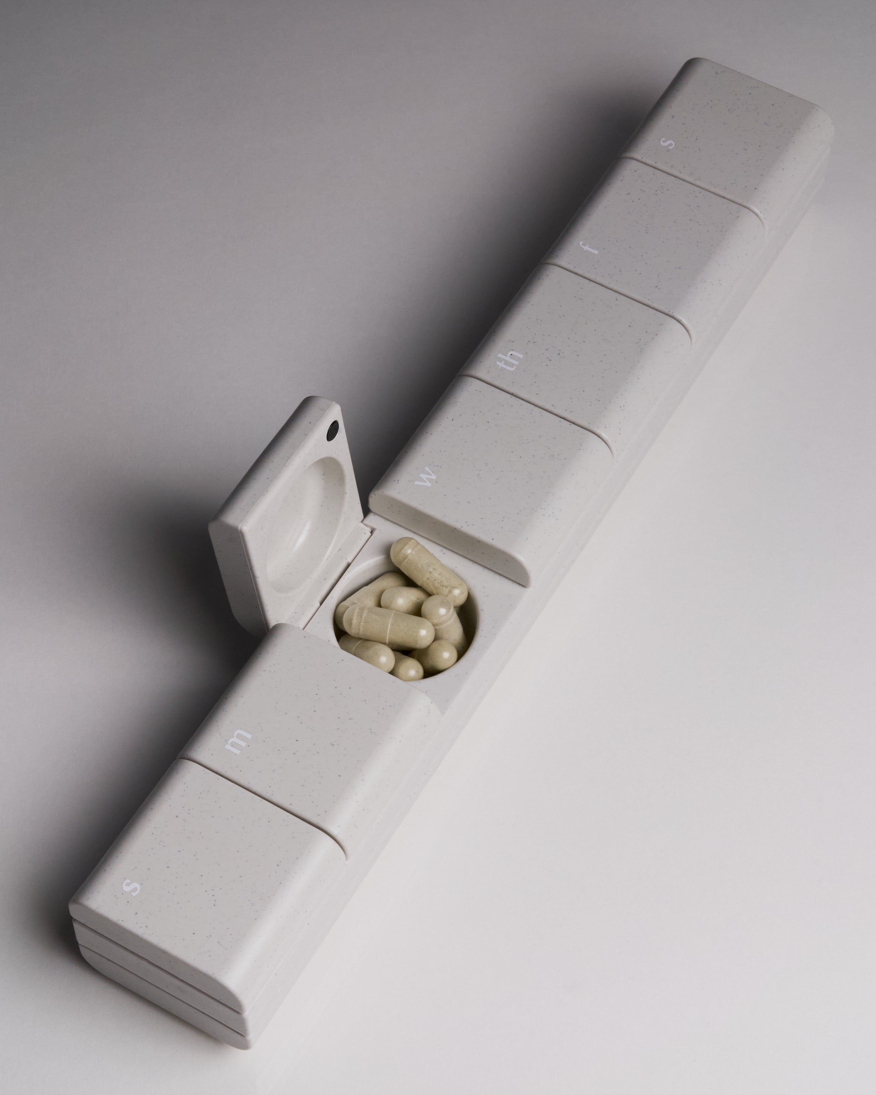 Top-down view of AndAgain 7-Day Pill Case in Original finish with one compartment open and capsules inside—minimalist design with weekday labels.