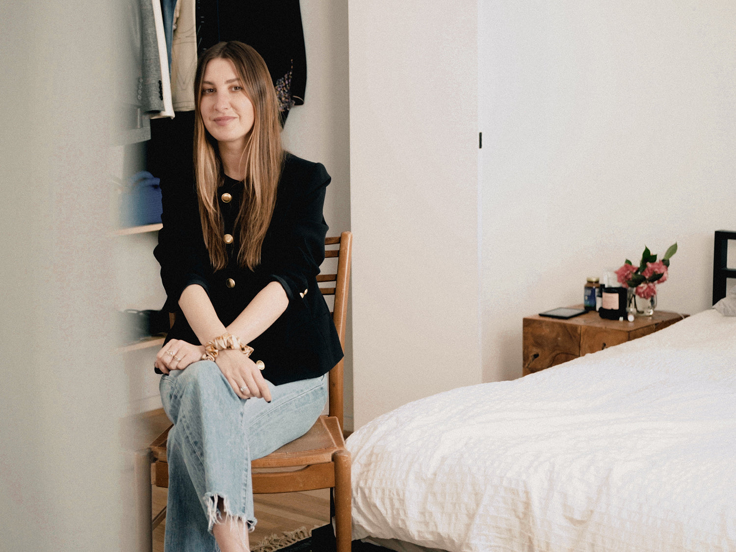 Allison Bornstein sitting on a wooden chair next to her bed, arms crossed, with a closet behind her, looking at the camera.