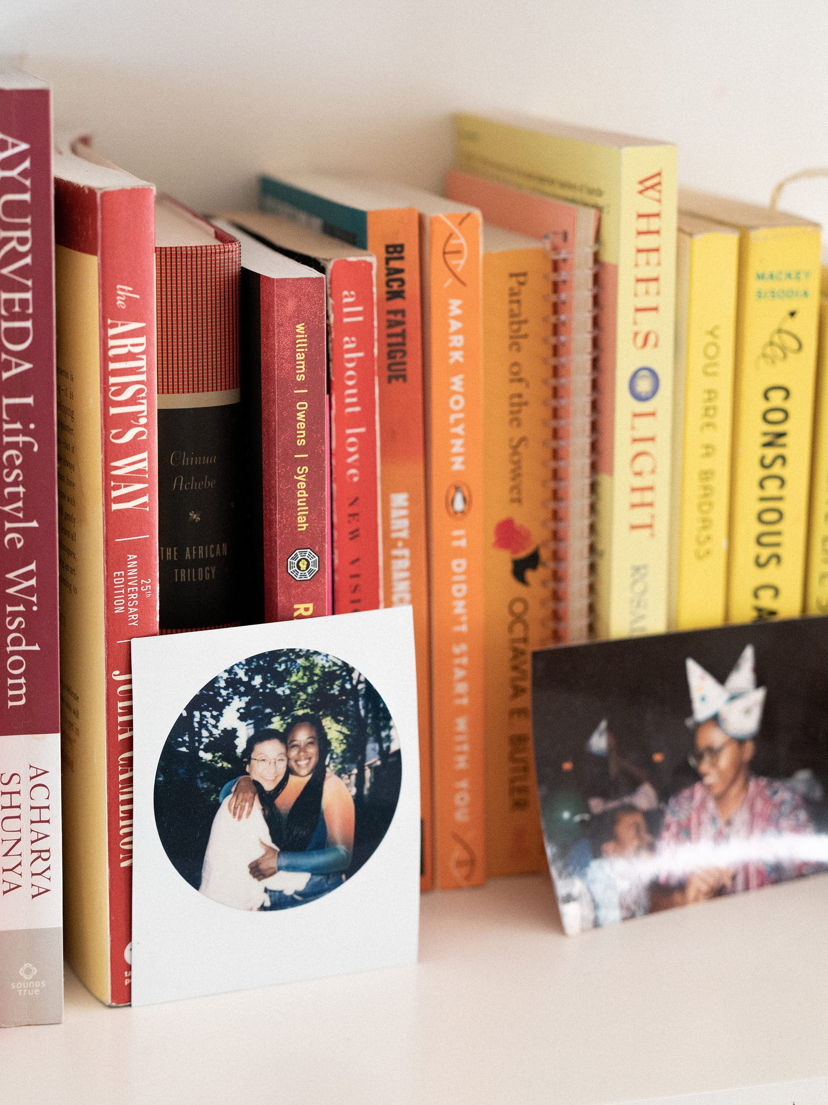 The founder of Dive in Well, Maryam Ajayi's book shelf with books aligned by color and a few printed photos leaning in front of them