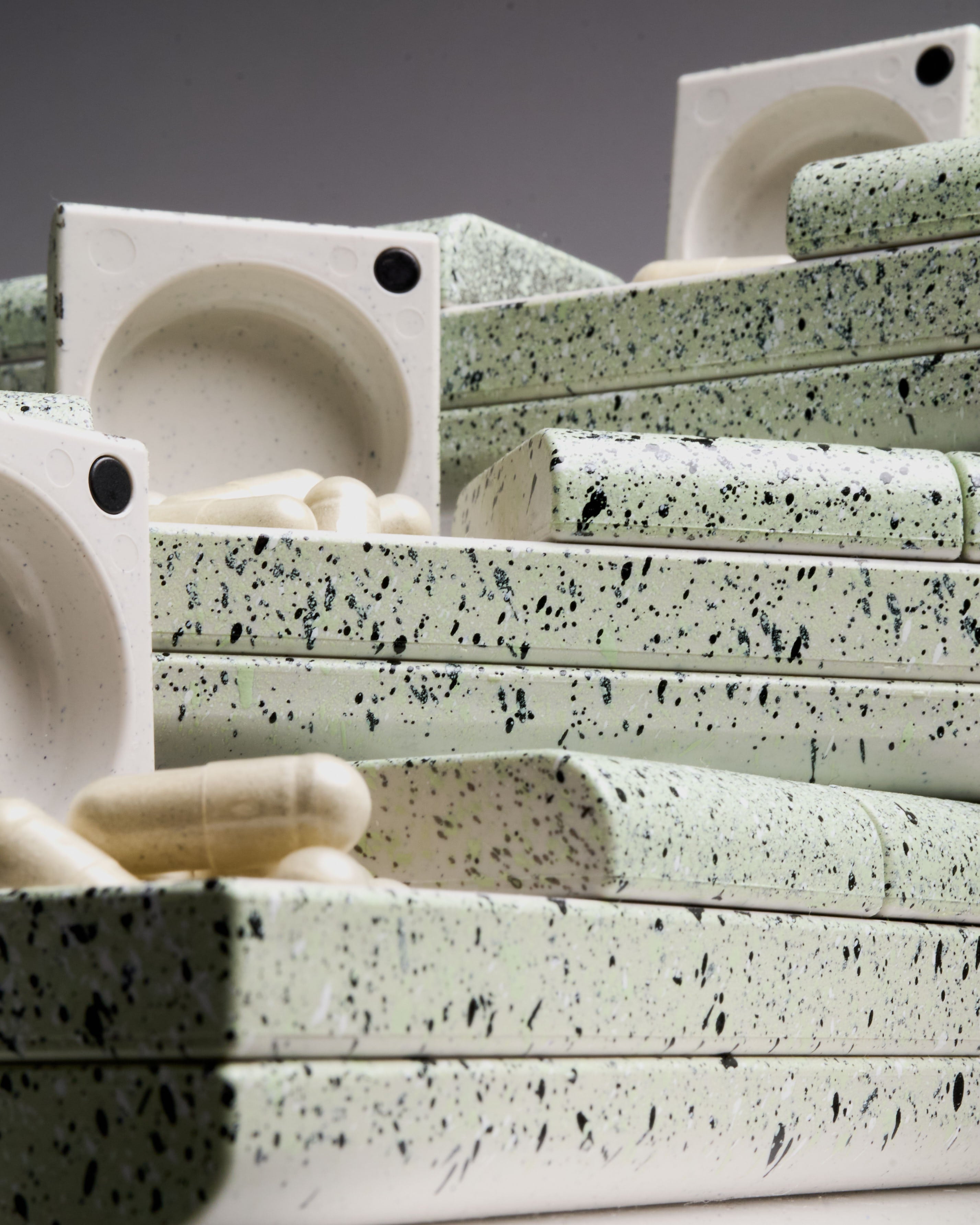 Stacked AndAgain Mint pill cases with open lids revealing capsules – speckled sage green pill organizers for modern supplement routines.